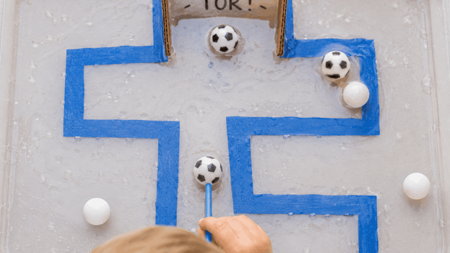 Kind pustet kleine schwimmende Bälle durch eine Wasserbahn zu einem Fußballtor. Kreatives Wasserspiel für Kinder mit Fußball-Thema.