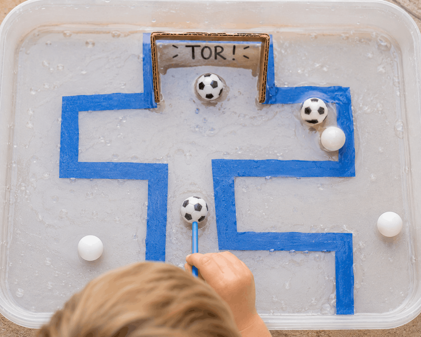 Kind pustet kleine schwimmende Bälle durch eine Wasserbahn zu einem Fußballtor. Kreatives Wasserspiel für Kinder mit Fußball-Thema.