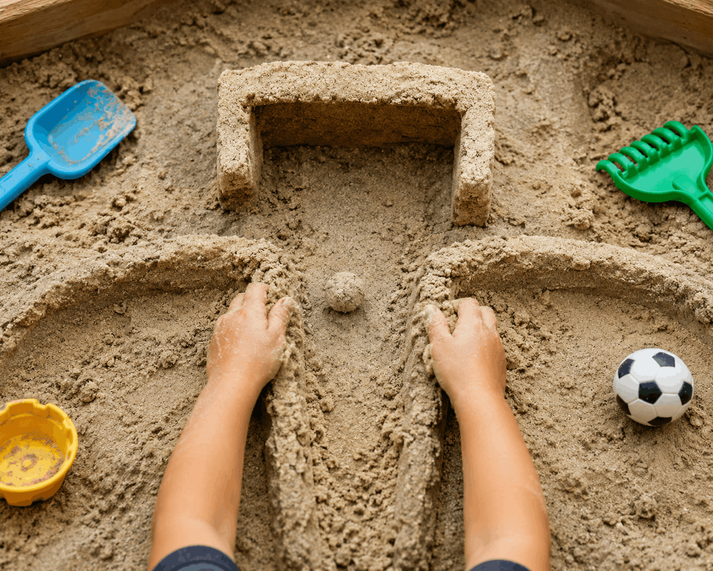 Kindliche Hände formen im Sandkasten ein kleines Fußballtor und bauen Wege für einen Ball. Sensorische Fußball-Spielidee mit Sand für Kinder.